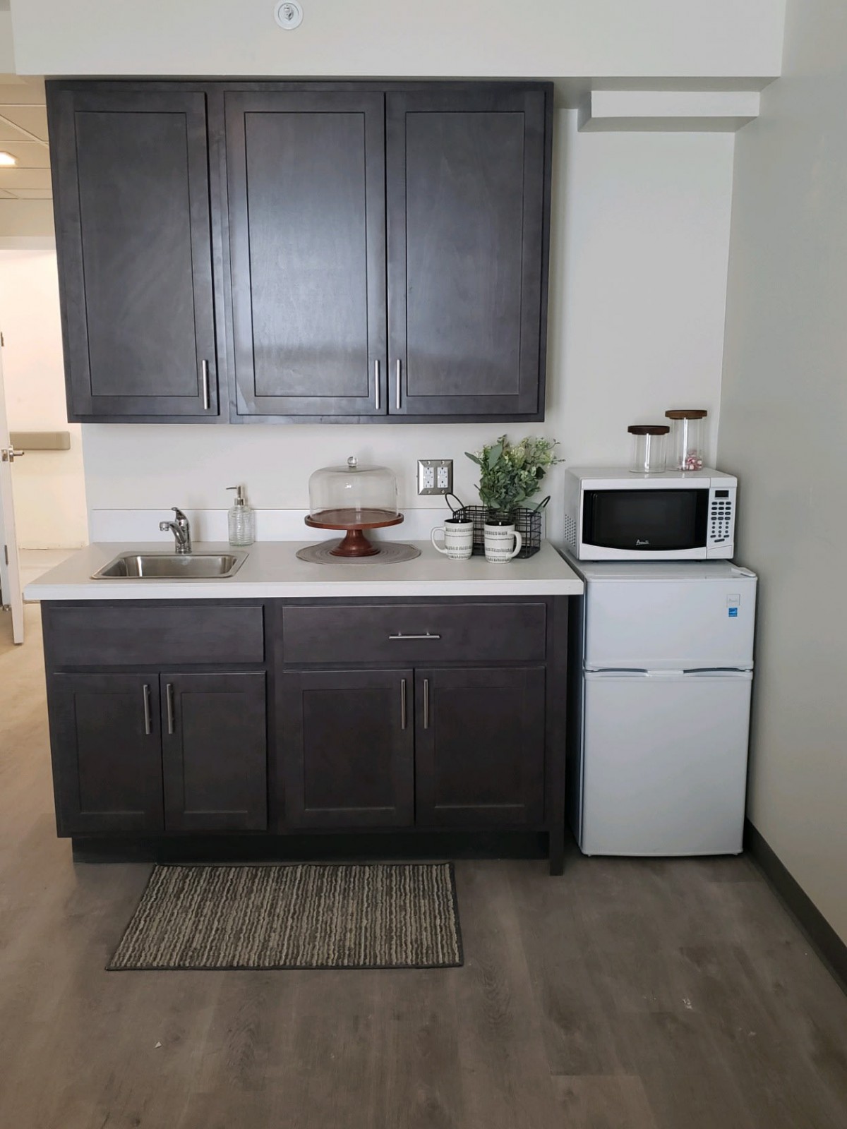 A compact kitchen area featuring dark cabinets, a sink, a microwave on top of a small refrigerator, and countertop decor.