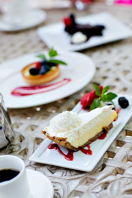 A beautifully plated dessert featuring a slice of cake with whipped cream, garnished with fresh berries and drizzled with raspberry sauce. A tart is visible in the background.