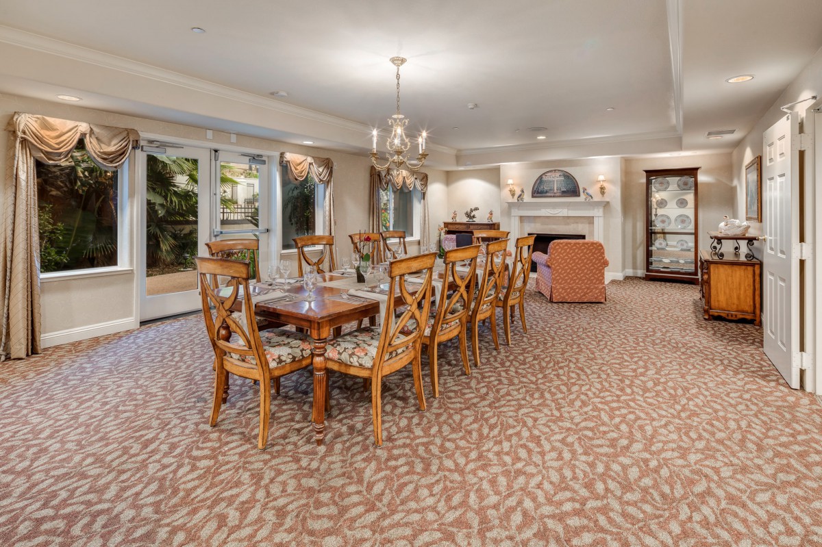 A spacious dining room featuring a long wooden table with matching chairs, adorned by an elegant chandelier and complemented by a patterned carpet.
