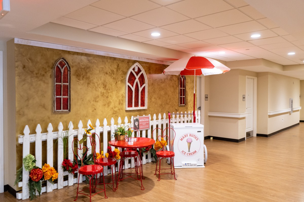 A cozy indoor setup with red tables and chairs by an artificial white picket fence, featuring decorative elements like an umbrella and wall art.