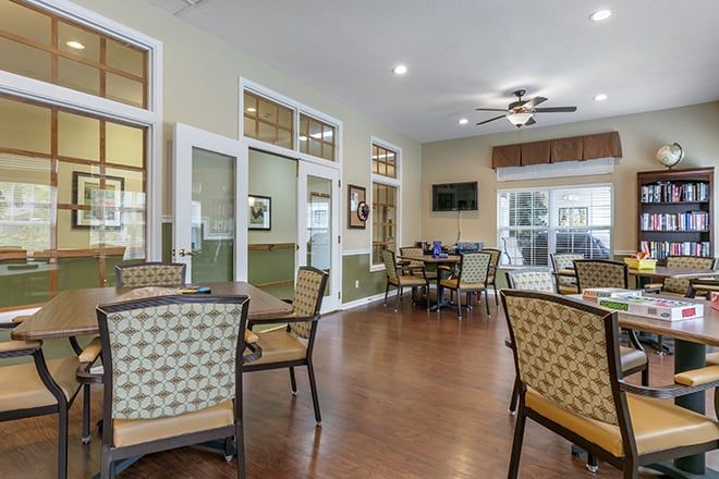 A cozy dining area with tables, chairs, and a ceiling fan, featuring large windows and a television.