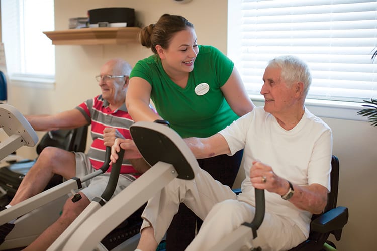A caregiver assisting elderly men with exercise using fitness equipment.