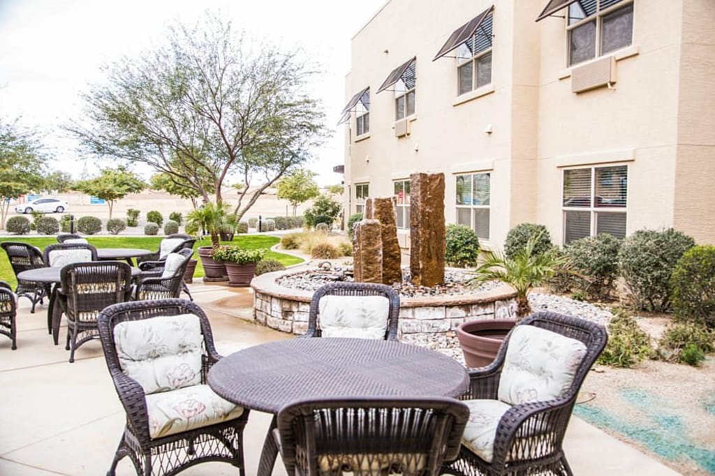 A cozy outdoor patio seating area with comfortable chairs and a stone water feature, set in a landscaped garden beside a building.