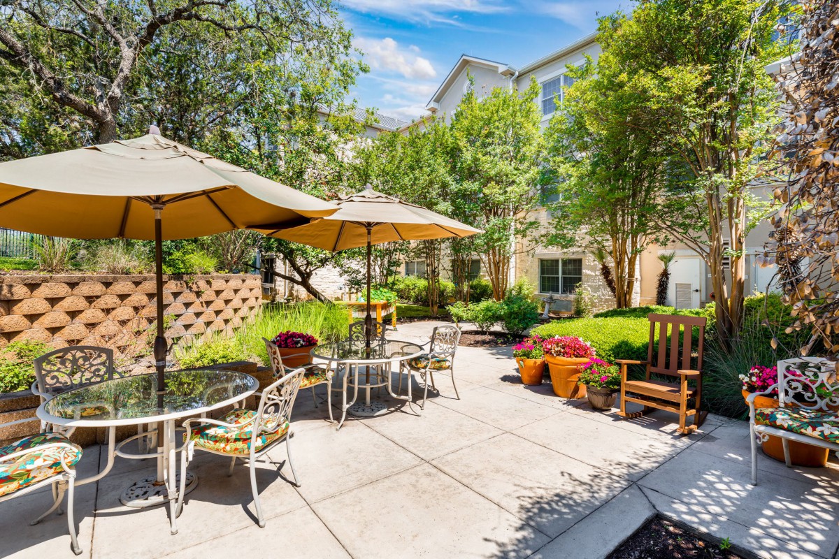 A sunny garden patio with tables and chairs under umbrellas, surrounded by lush greenery.