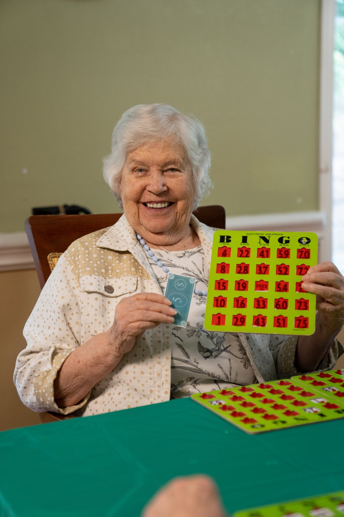 An elderly woman joyfully holding a bingo card while playing the game indoors.