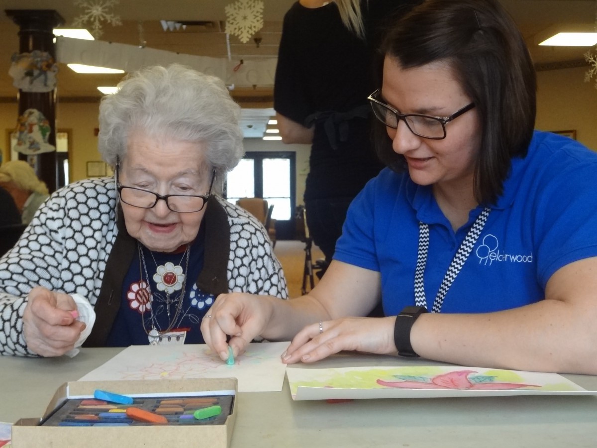 A young woman assists an elderly woman with a coloring activity indoors.
