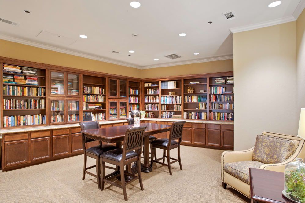 A cozy library room with wooden bookshelves filled with books, a central table surrounded by chairs, and warm lighting.
