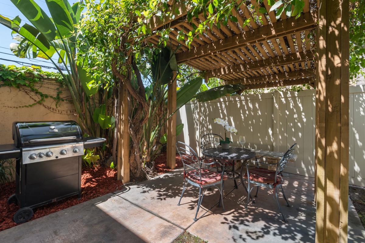 A cozy garden patio with a pergola, outdoor seating, and a grill surrounded by lush plants.