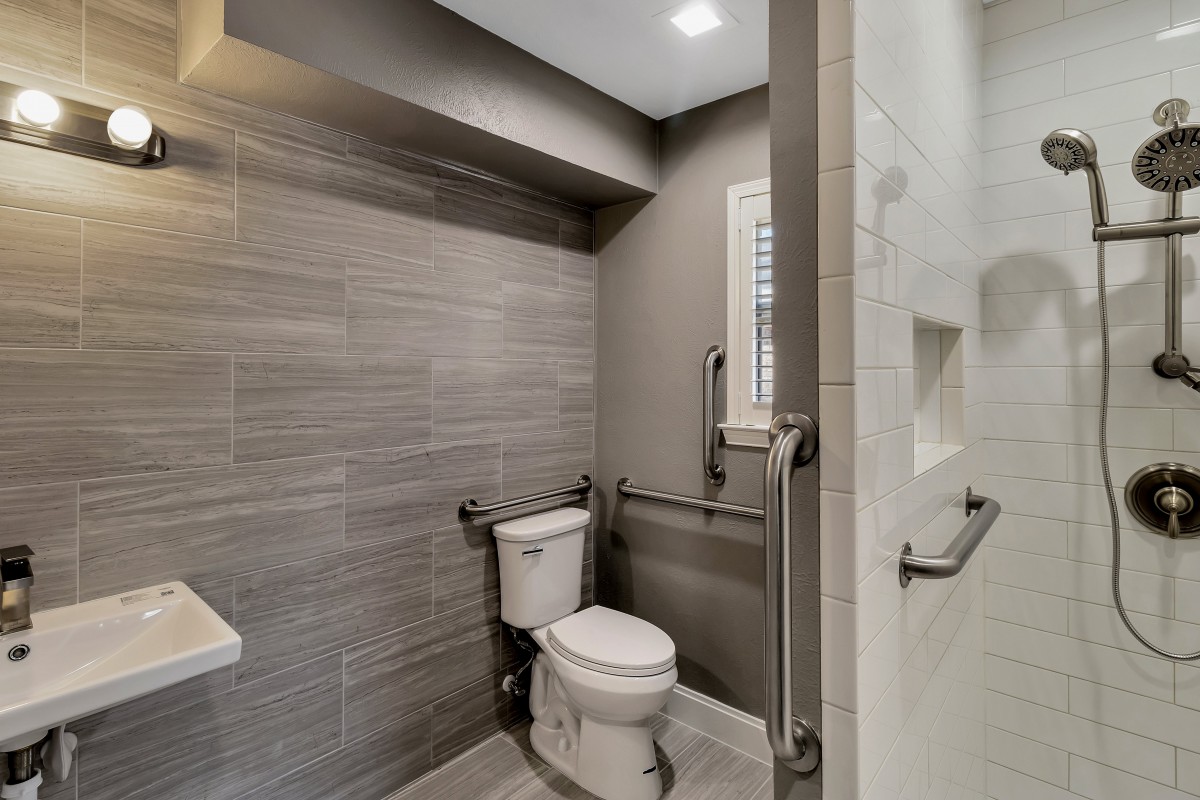 A modern bathroom featuring gray tiles, a toilet with grab bars, and a shower area.