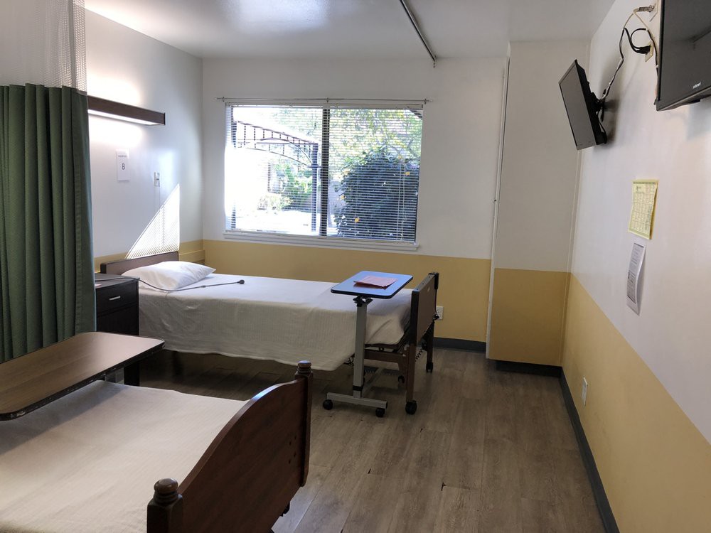 A simple hospital room with a single bed, a window, and a wall-mounted television.