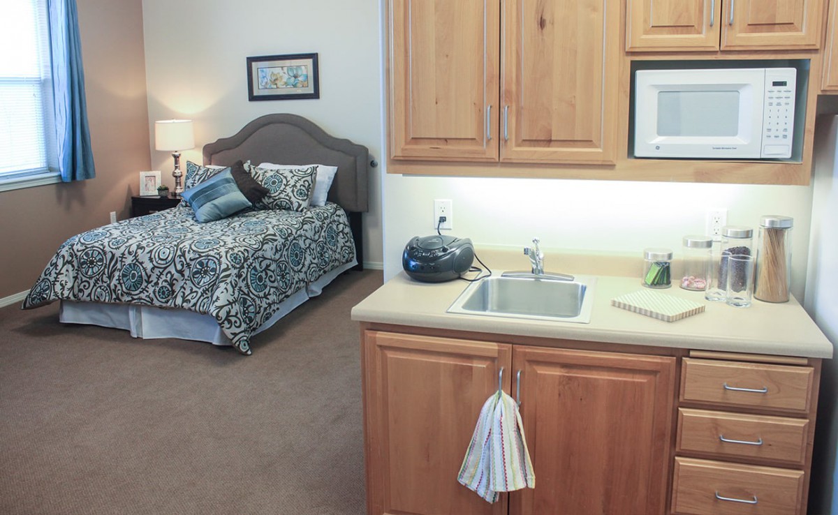 A cozy bedroom with a small kitchenette, featuring a bed, lamp, and sink.