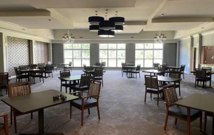 An empty dining room with neatly arranged tables and chairs, featuring large windows and elegant chandeliers.
