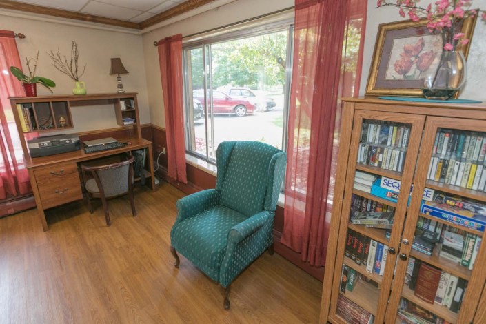 A cozy living room corner with an armchair by the window, a bookshelf, and a small desk.