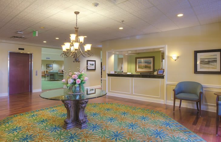 A brightly lit hotel lobby with a reception desk and a floral arrangement on a round table.