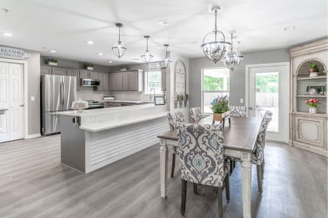 A modern open-concept kitchen and dining area with sleek gray tones, featuring chandeliers, stainless steel appliances, and elegant patterned chairs.