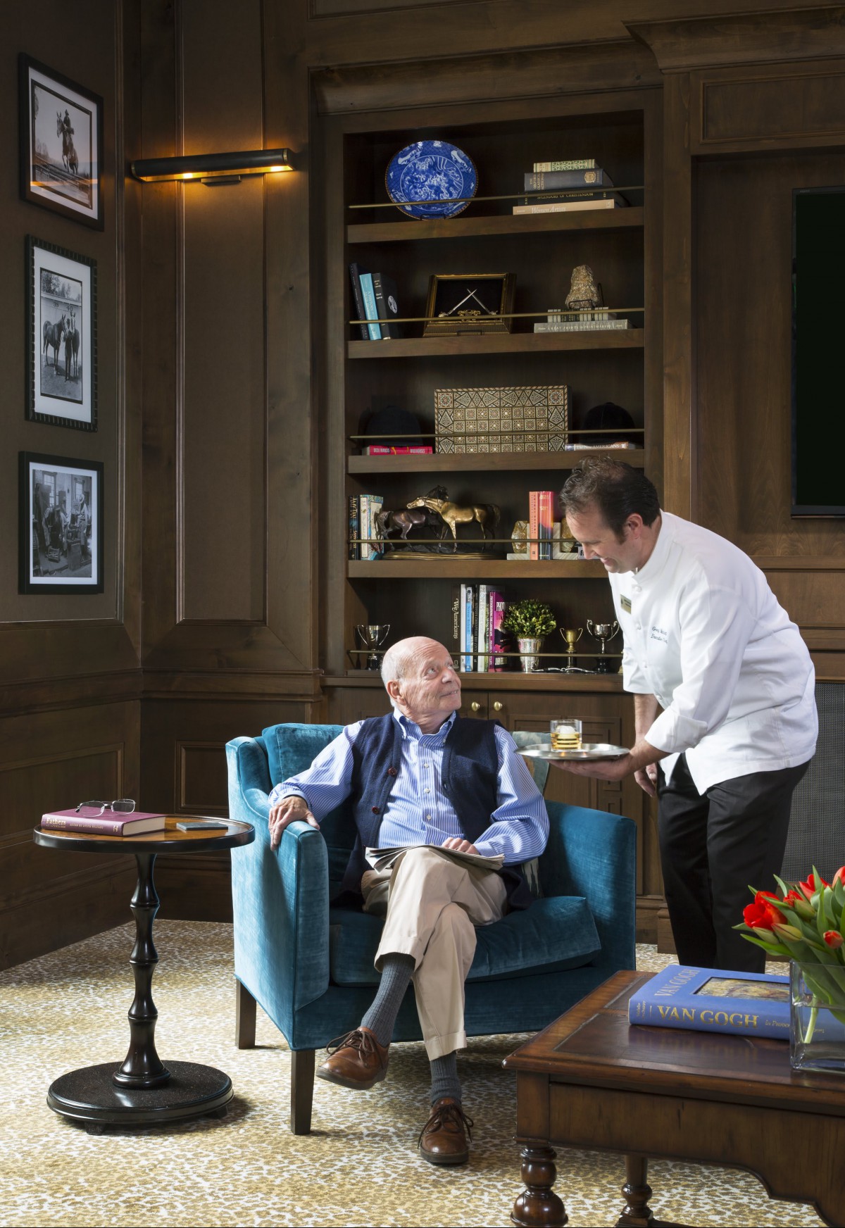 An elderly man sitting in an armchair in a library is being served by a waiter.