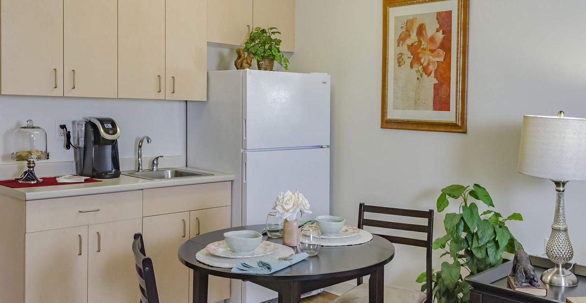A cozy kitchen with a dining table set for two, featuring a refrigerator, plants, and wall artwork.
