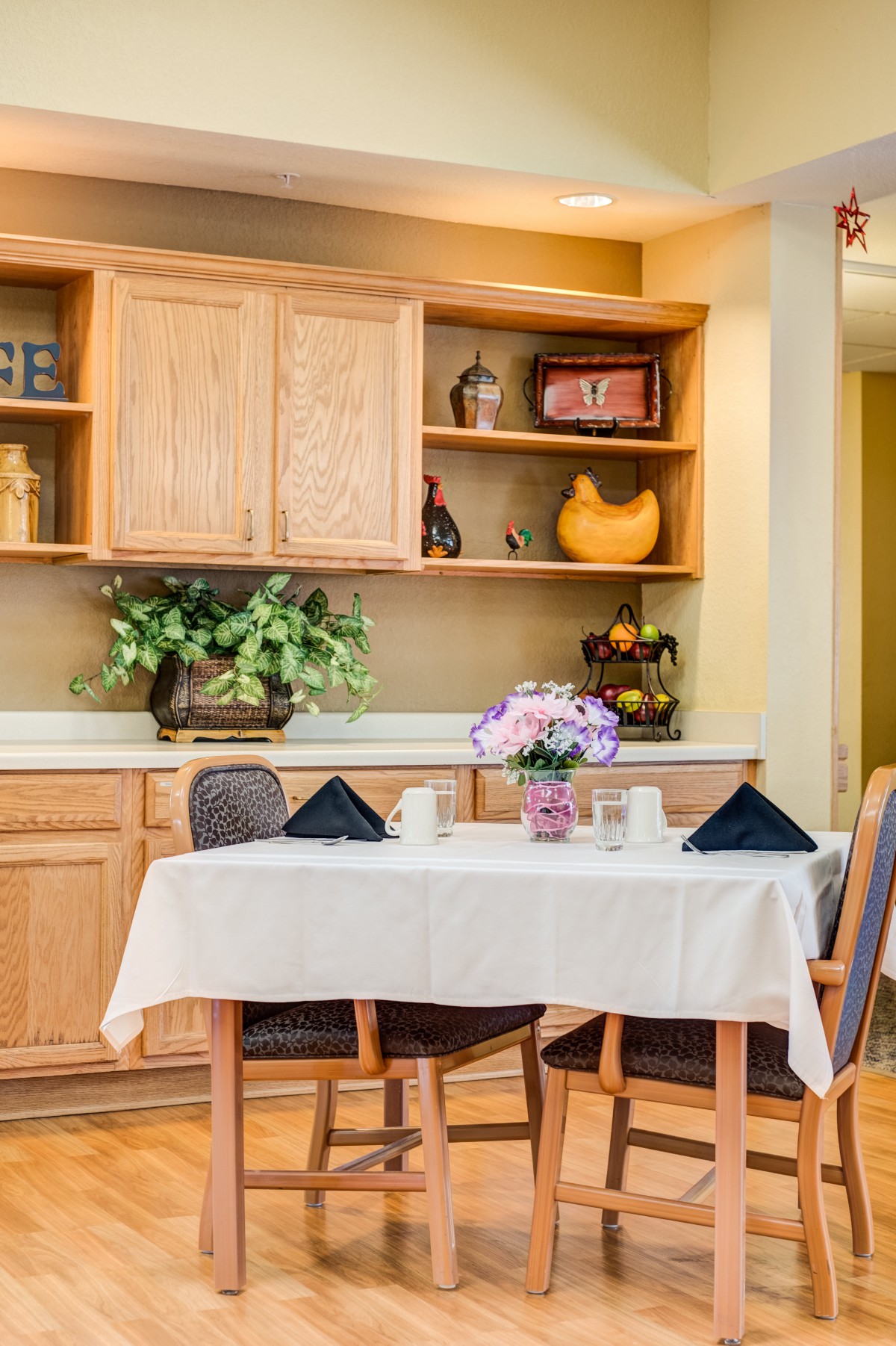 A cozy dining area with a table set for two, featuring a floral centerpiece and surrounded by wooden cabinets.