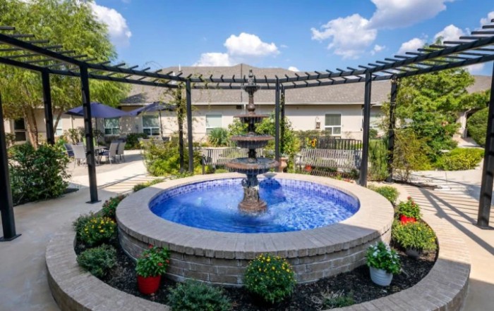 A serene garden setting featuring a central fountain surrounded by a pergola and lush greenery.
