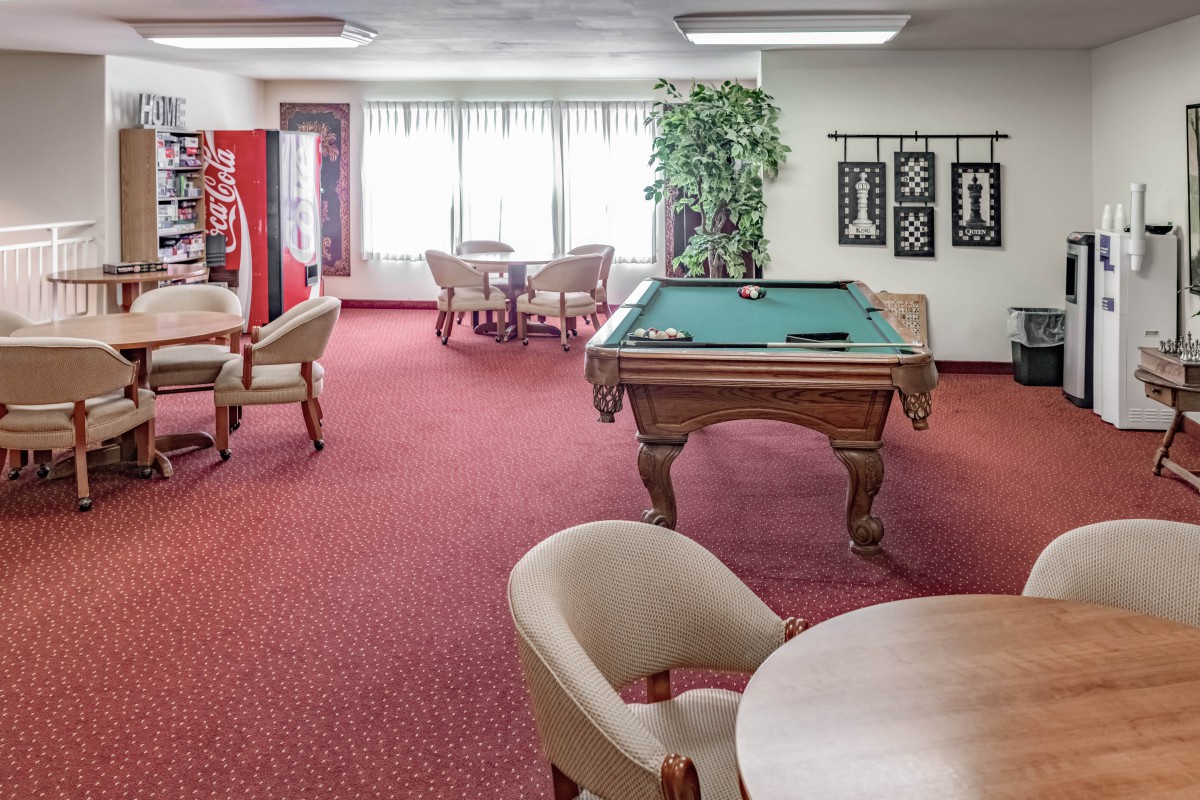 A recreational space featuring a pool table, several tables and chairs, a vending machine, and an indoor plant on a carpeted floor.
