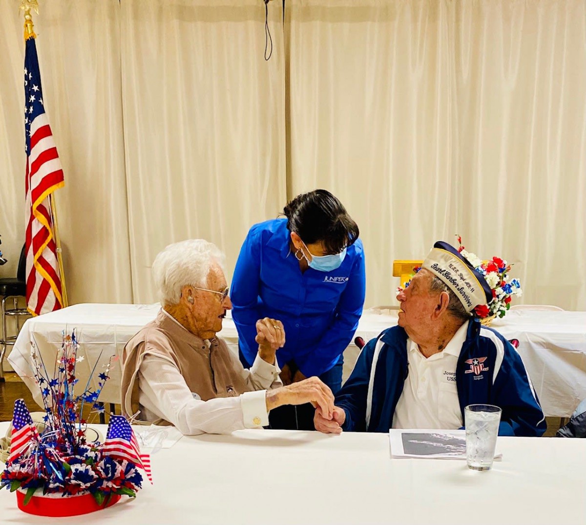 Two elderly individuals, possibly veterans, engage in conversation at a celebration event adorned with patriotic decorations, including an American flag.