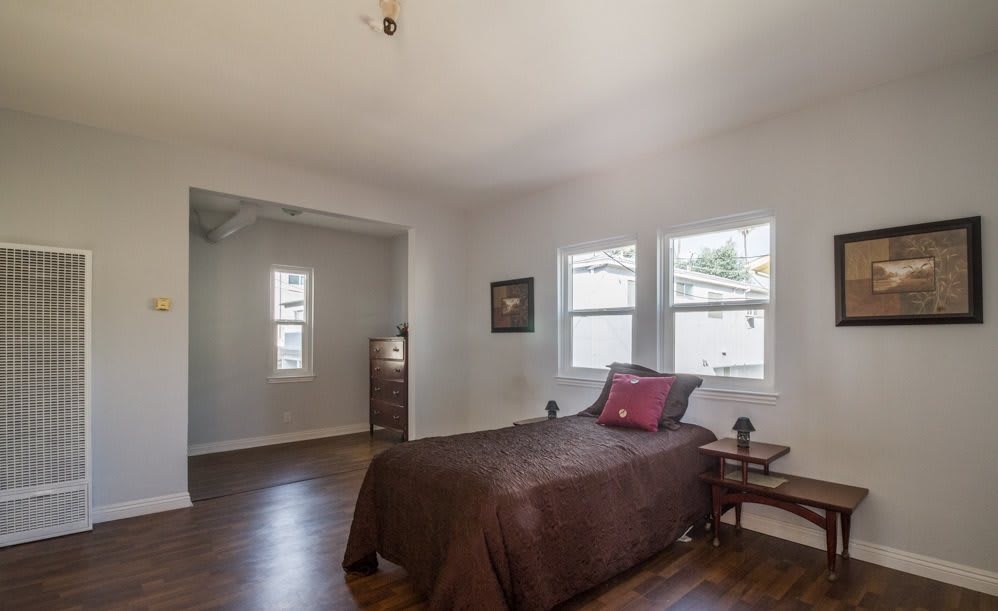 A cozy bedroom with a single bed, wooden floor, and natural light coming through the windows.