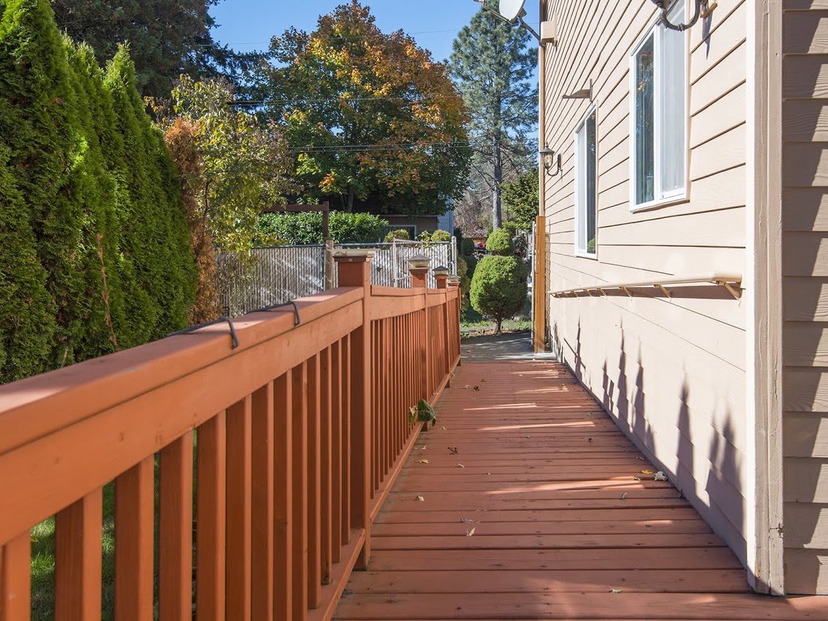 A sunny view of a wooden deck alongside a house, bordered by a fence and surrounded by trees.