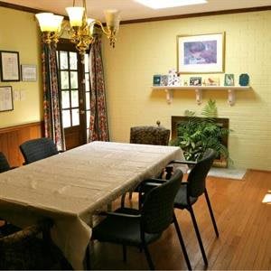 A cozy meeting room with a rectangular table, surrounded by black chairs. The room features a chandelier, a fireplace with decorative items, framed artwork on the walls, and a potted plant for added ambiance.