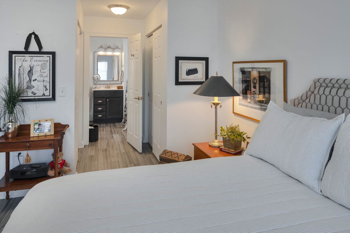 A modern bedroom featuring a neatly made bed, bedside table with a lamp, and framed artwork on the wall. The view extends into a hallway leading to a bathroom.