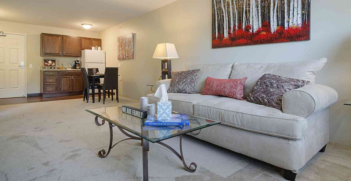 A cozy living room with a glass coffee table, comfortable sofa, and warm lighting leading to a dining area.