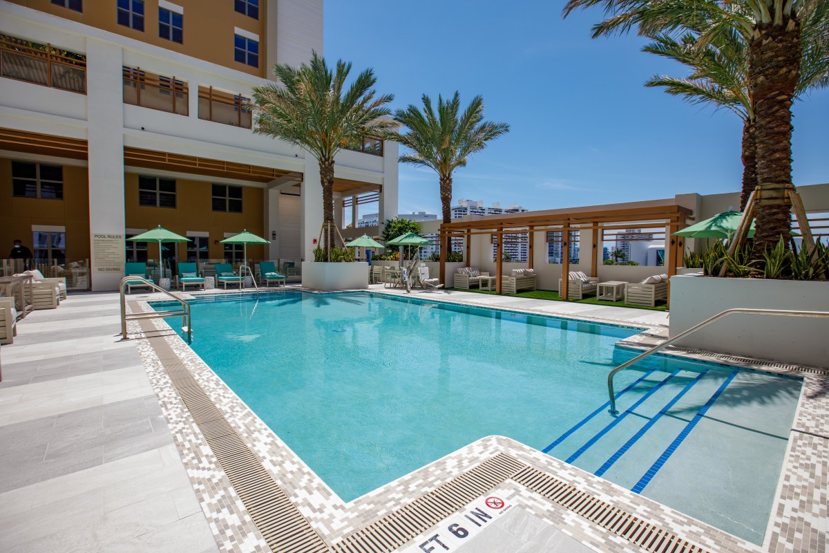Outdoor hotel swimming pool area with lounge chairs and palm trees on a sunny day.