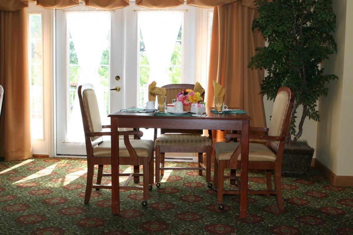 A well-lit dining area featuring a wooden table and four chairs, with large windows and curtains providing natural light.