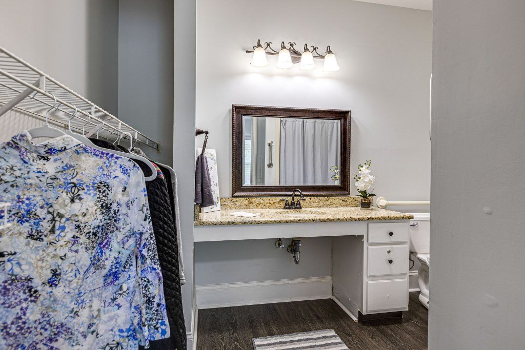 A bathroom vanity with a mirror and lighting, adjacent to a clothing rack.