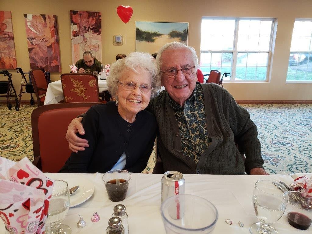 An elderly couple smiling and sitting together at a dining table indoors.