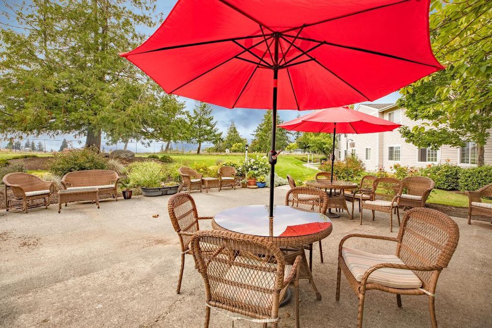A peaceful patio with wicker chairs and tables under red umbrellas, overlooking a scenic garden.