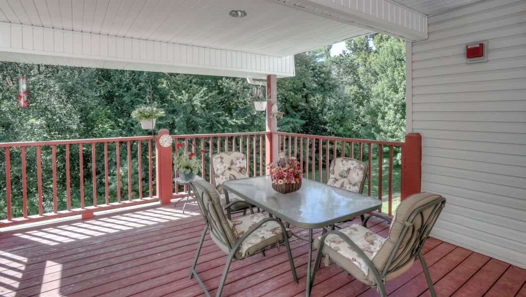 A cozy covered deck with a glass table and four cushioned chairs, surrounded by wooden railings and overlooking lush greenery.