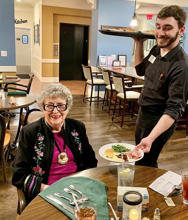 A senior woman sits at a table in a restaurant while a waiter serves her meal.