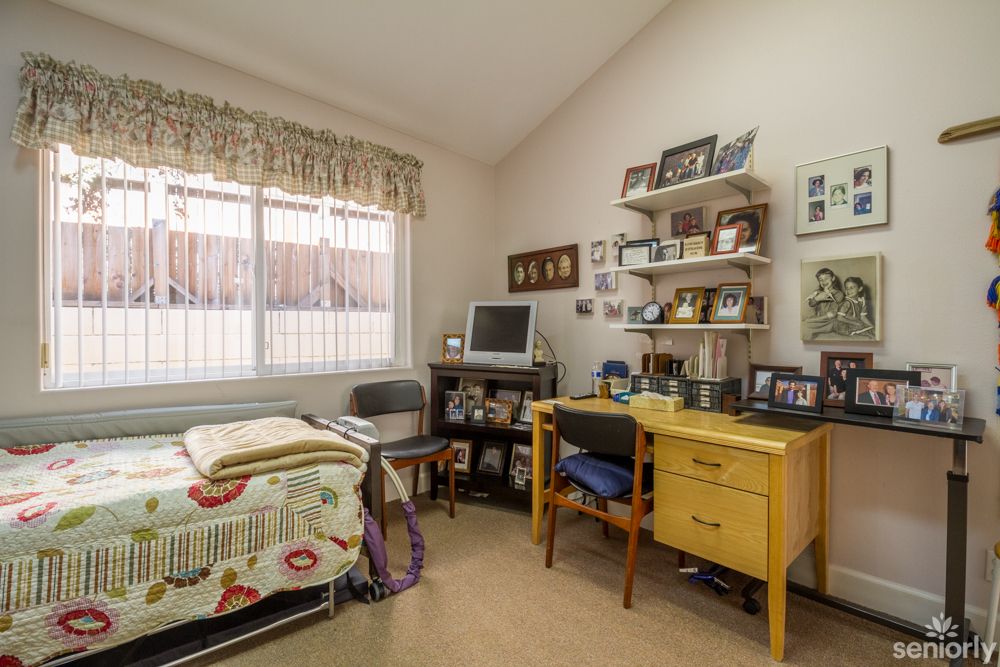 A cozy bedroom with a small workspace, featuring a desk and shelves filled with framed pictures.