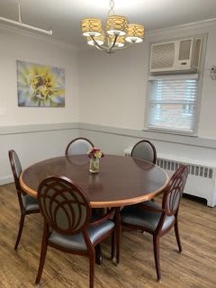 A small dining room with a round table and four chairs, featuring a wall painting and window with an air conditioning unit.