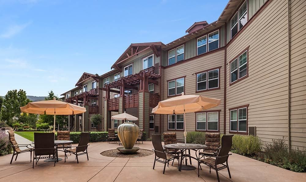A view of a residential building with a patio area featuring tables, chairs, umbrellas, and a fountain.