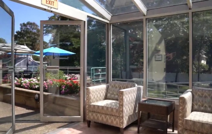 A cozy lounge area with a patterned chair, surrounded by large windows showcasing a sunny outdoor patio.