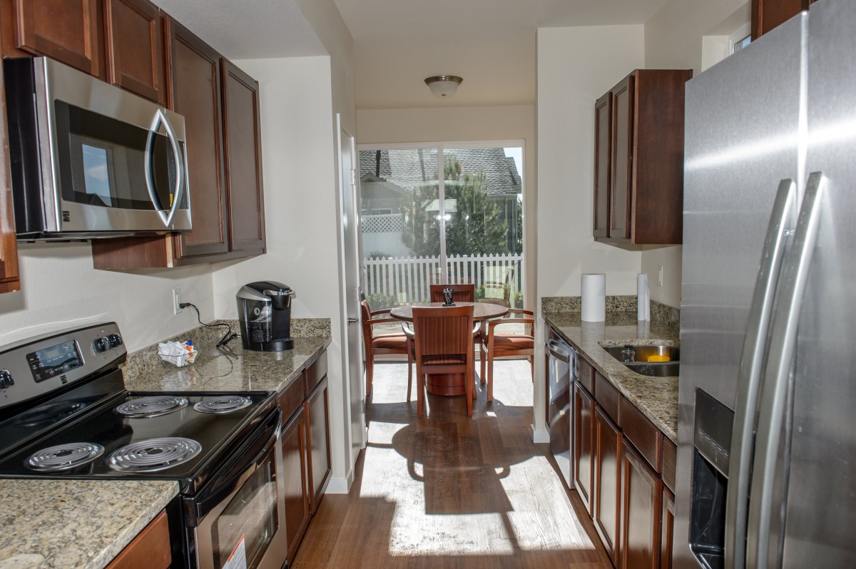 A modern kitchen with stainless steel appliances and wood cabinets opens into a sunlit dining area.