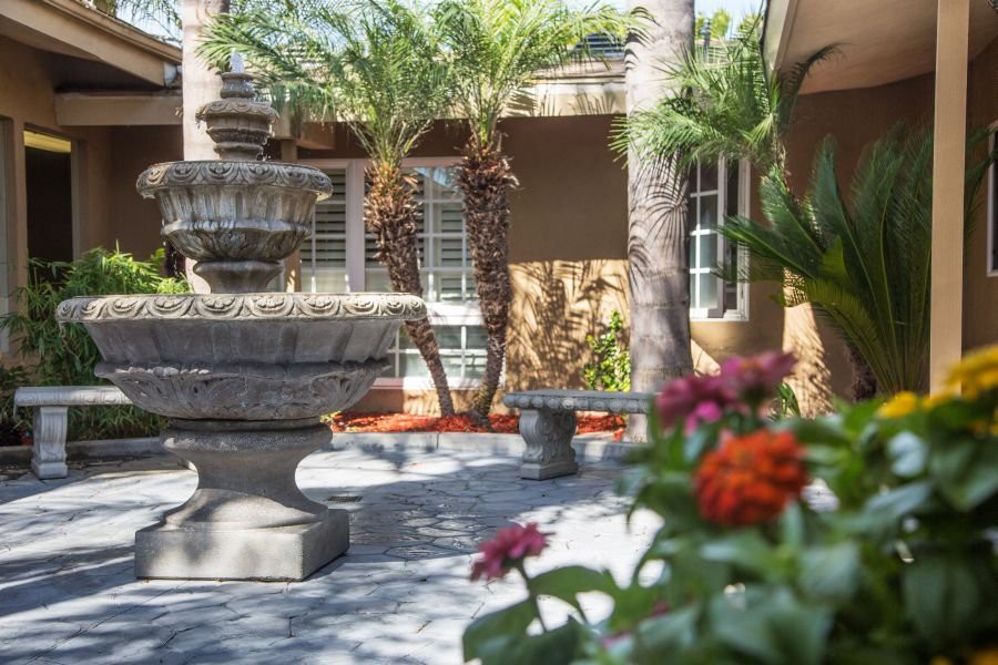 A tranquil courtyard featuring a stone fountain surrounded by palm trees and colorful flowers.