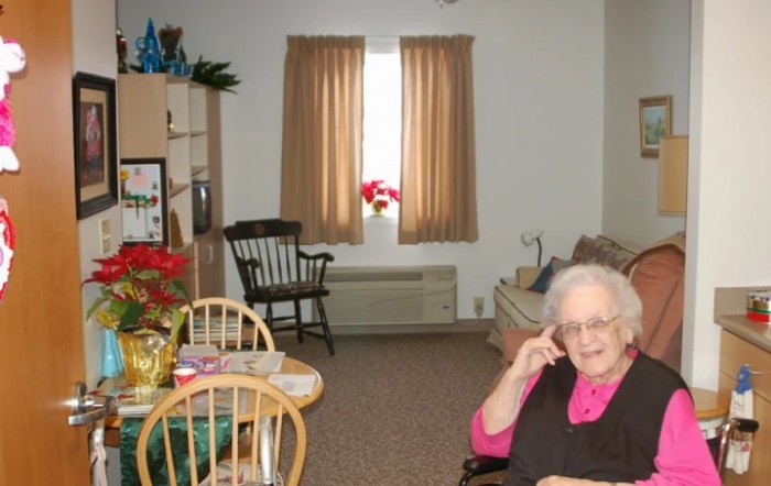A senior woman sits in a cozy living room with simple furnishings.