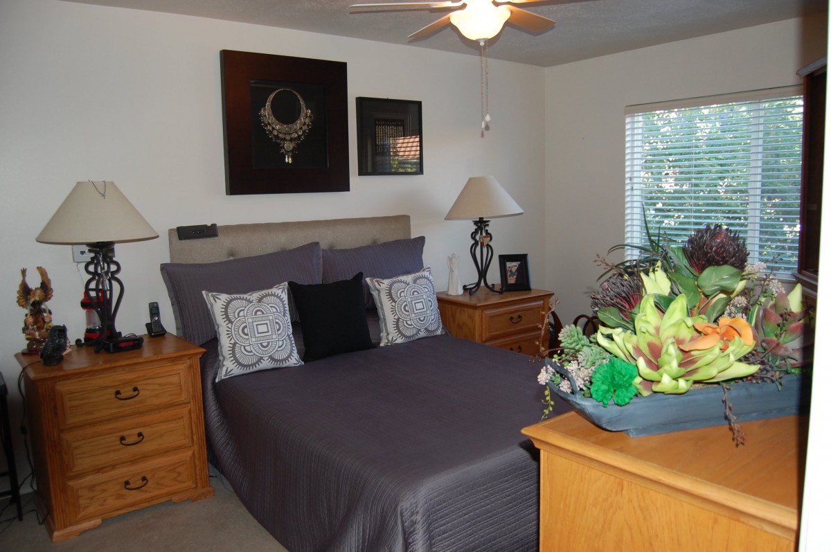 A cozy bedroom featuring a neatly made bed with dark bedding, wooden nightstands, decorative wall art, and indoor plants.