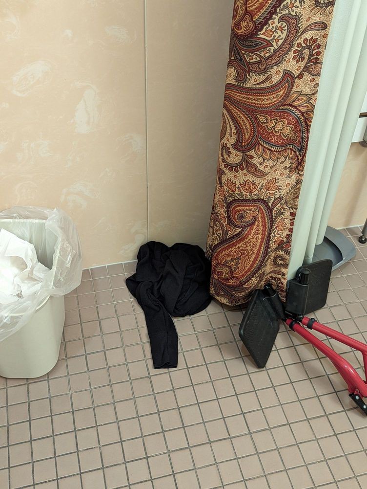 A cluttered corner of a bathroom with a coat, trash can, carpet, and exercise equipment.