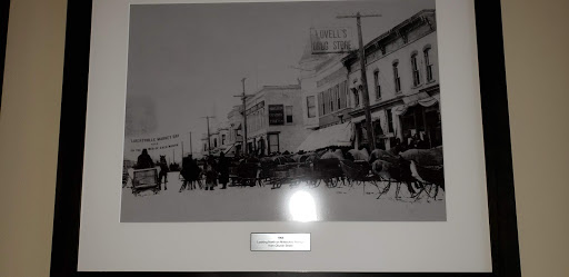 A historical black and white photograph depicting a busy street scene with horse-drawn carriages and pedestrians in front of early 20th-century buildings.