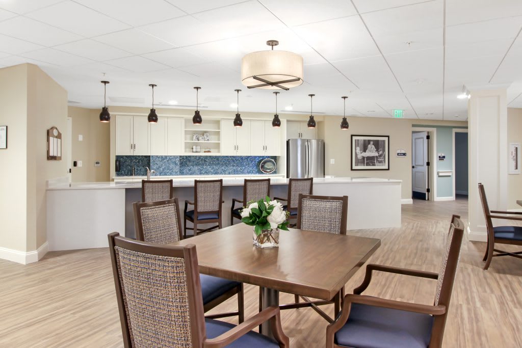 A modern dining area with a wooden table and upholstered chairs, adjacent to a spacious kitchen featuring pendant lights.
