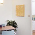 A dining area with a wooden table, blue chairs, a potted plant, and a piece of yellow artwork on the wall.