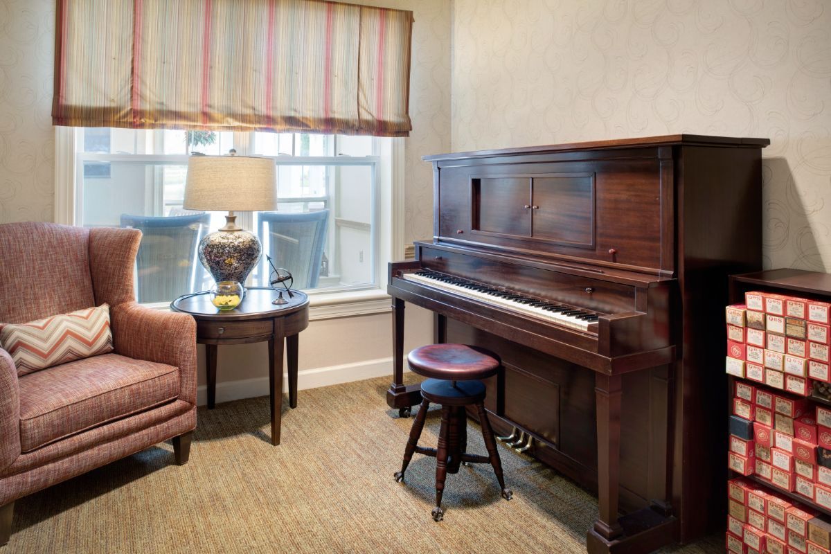 A cozy music room featuring an upright piano, a stool, and a side table with a lamp next to a window.